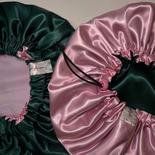 Two reversible satin bonnets, one displayed in sage green and the other in soft pink, both with drawstrings and elasticated bands.