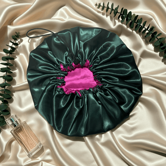 Green satin hair bonnet with pink interior on a beige fabric background with a perfume bottle and leaves.