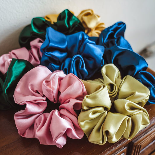 A photo of a variety of satin scrunchies in pink, blue, green, and yellow. They are placed on a wooden surface. In the background, there is a white wall.