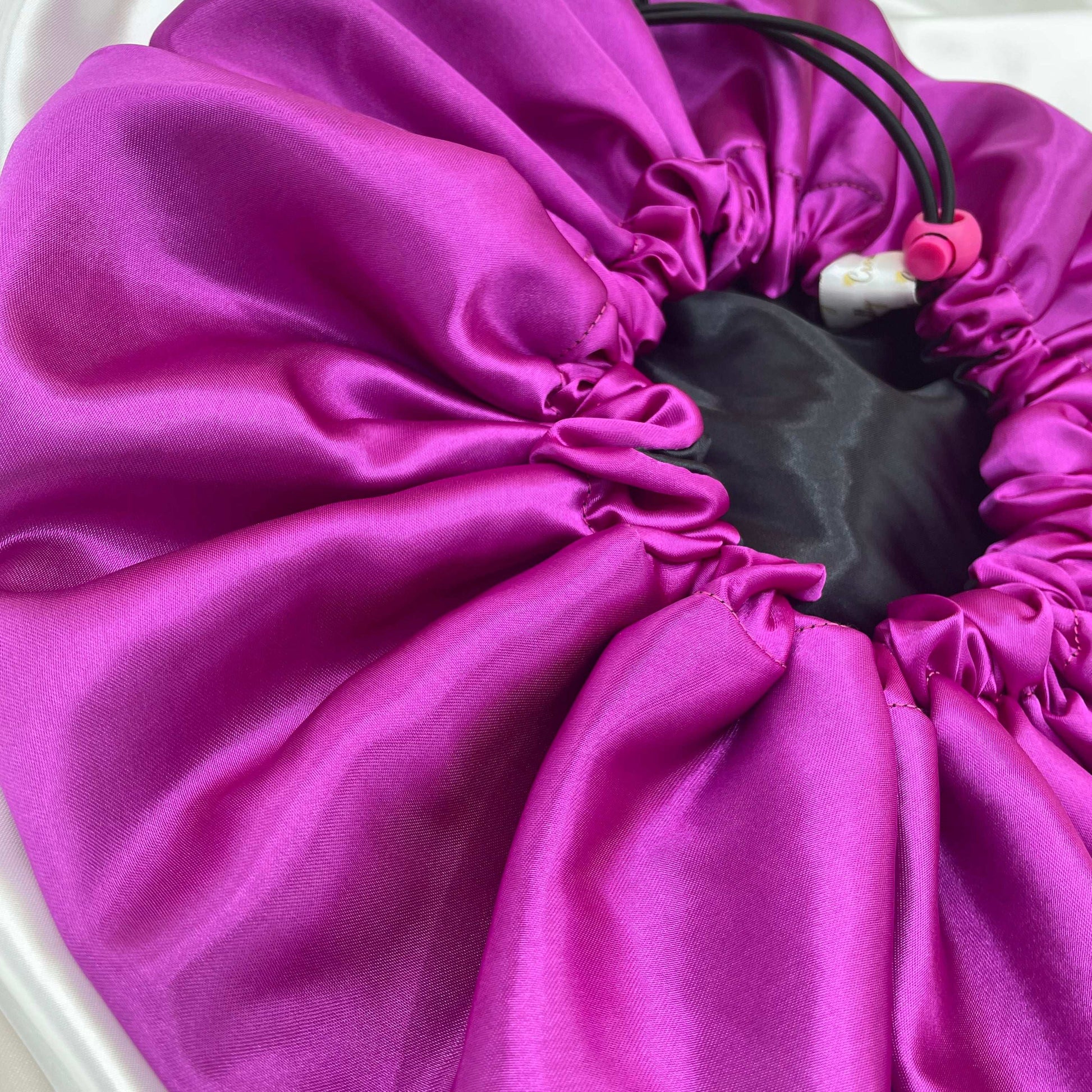 A close-up view of a reversible satin bonnet in magenta and black colors, showcasing the adjustable drawstring and elastic band.