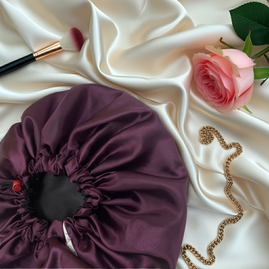 Purple satin bonnet on a beige fabric background with a rose, lip gloss, and gold chain.